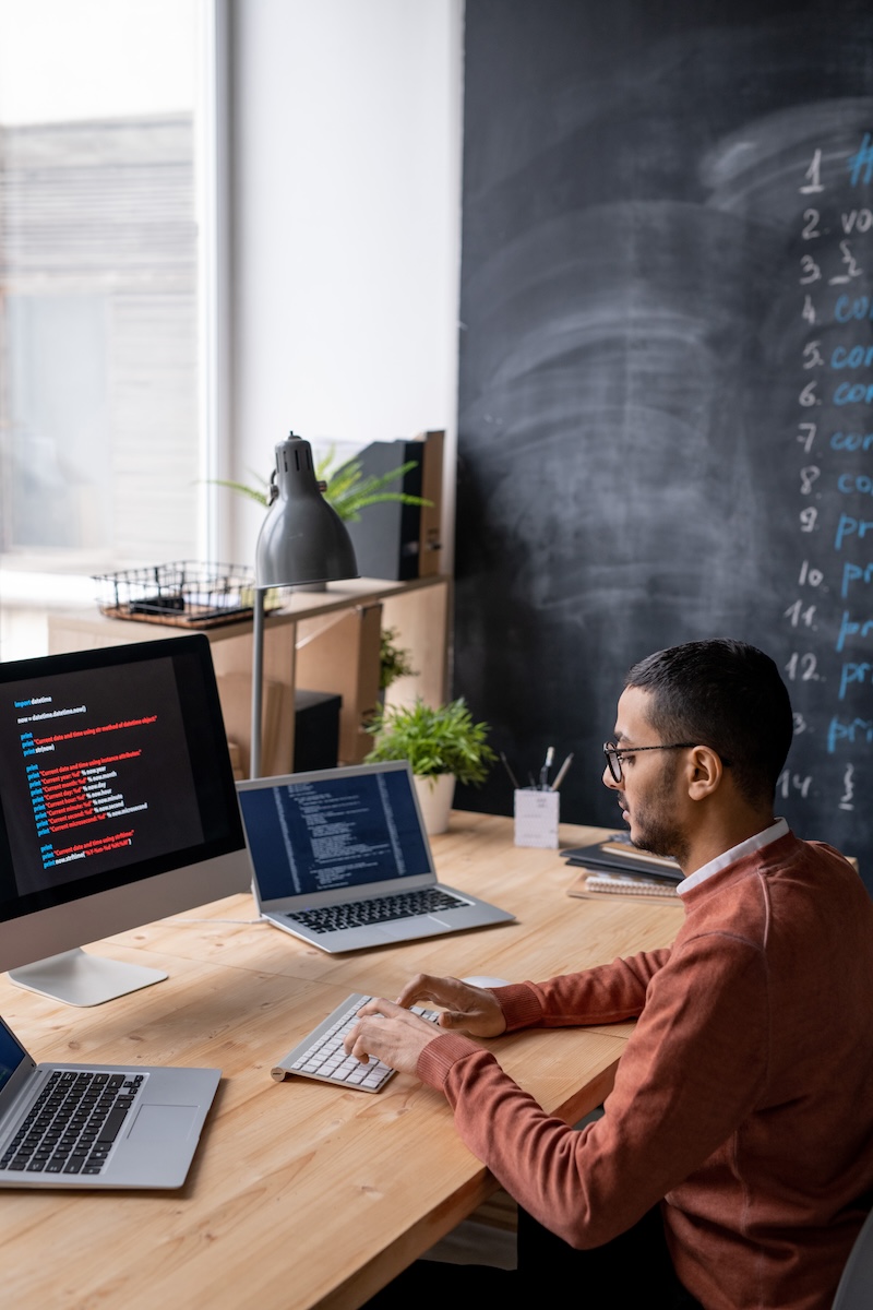 A professional working on a computer, displaying code on the screen, in a modern office setup with a chalkboard and potted plants in the background.